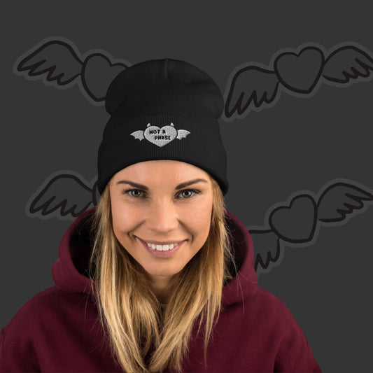 Person wearing an embroidered beanie with a winged a horned heart and text "not a phase"
