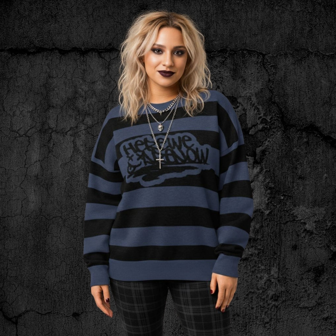 Alt style person wearing a blue and black grunge striped knitted sweater with text "here we are now" in graffiti tag style, against a dark textured background