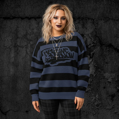 Alt style person wearing a blue and black grunge striped knitted sweater with text "here we are now" in graffiti tag style, against a dark textured background