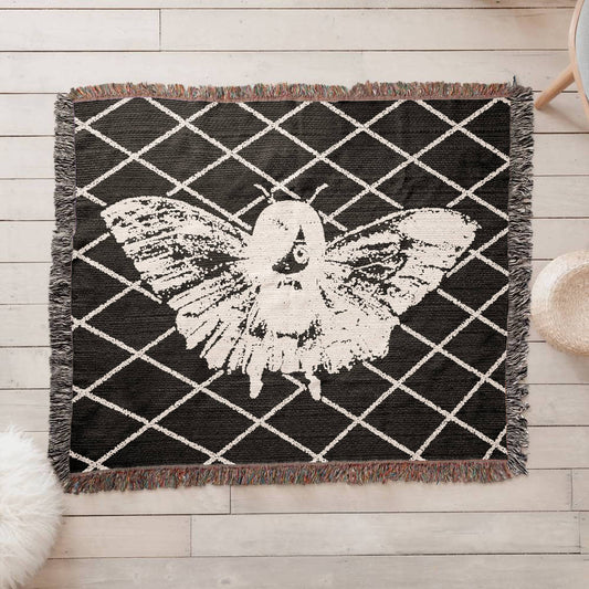 a woven blanket featuring a moth girl, wings spread on a background of a lattice grid on a wooden floor