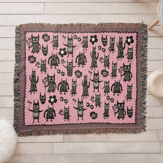 a pink woven blanket featuring grinning goon-goblins and flowers on a floor