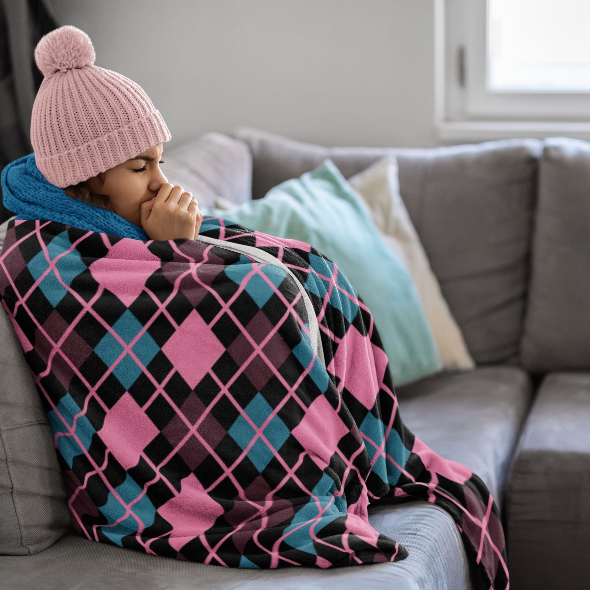 Person wrapped in a colorful argyle pattern blanket on a couch, wearing a pink knit hat.