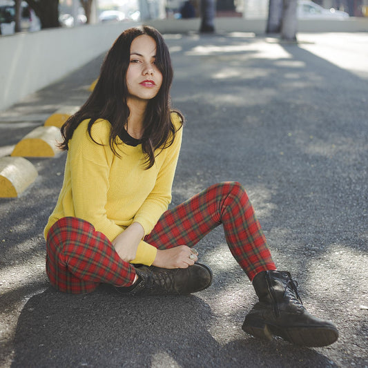 Woman sitting on a street wearing a yellow sweater and red plaid punk pattern leggings and boots.