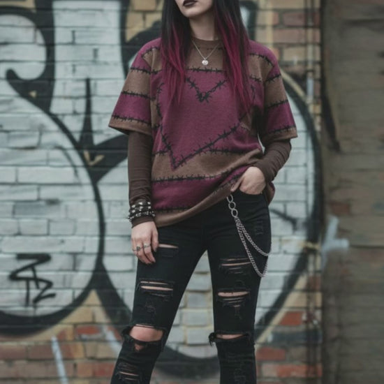 An alt style girl in a street setting wearing a knitted tee with dark barbed burgundy-brown stripes and a large barbed heart, worn over a long sleeve