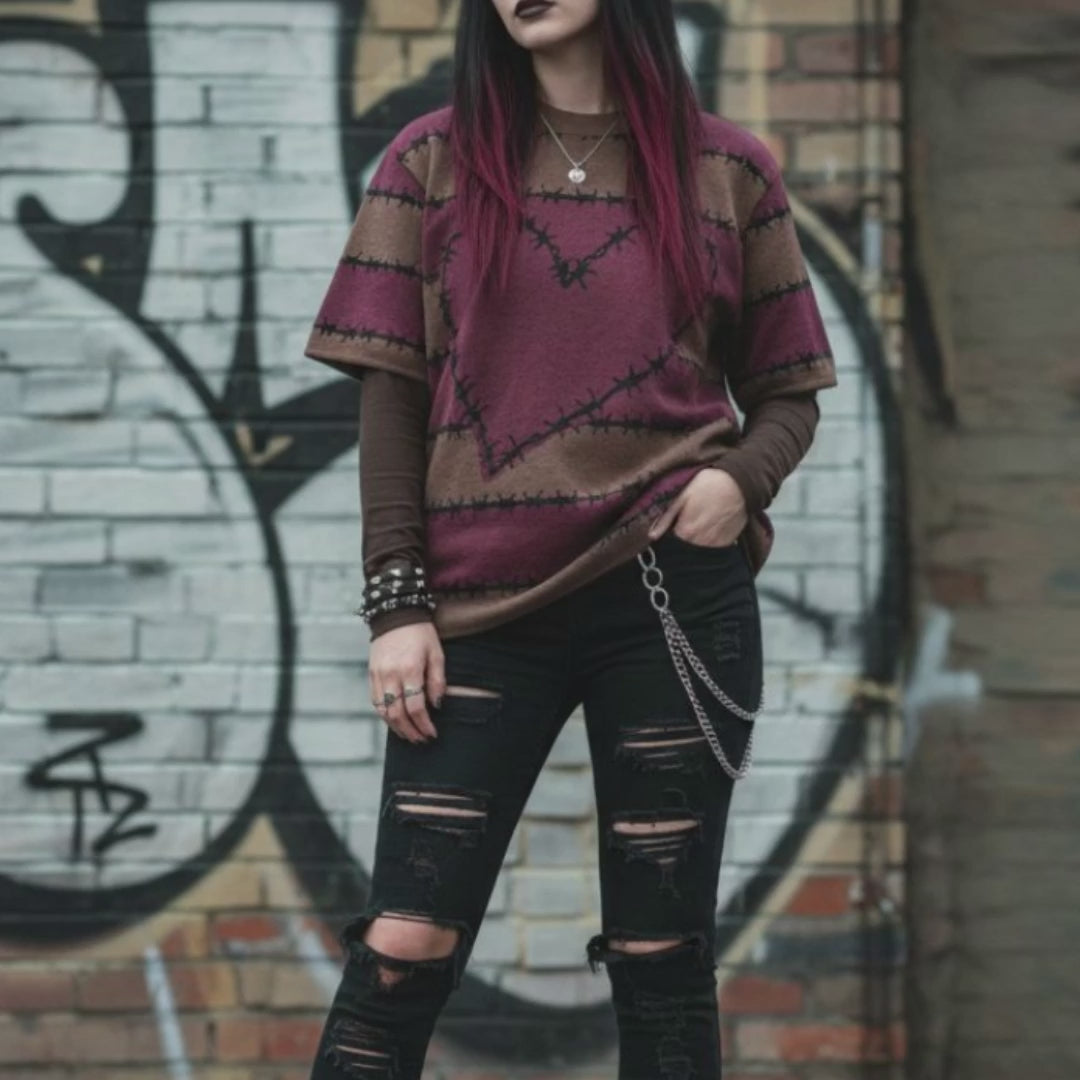 An alt style girl in a street setting wearing a knitted tee with dark barbed burgundy-brown stripes and a large barbed heart, worn over a long sleeve