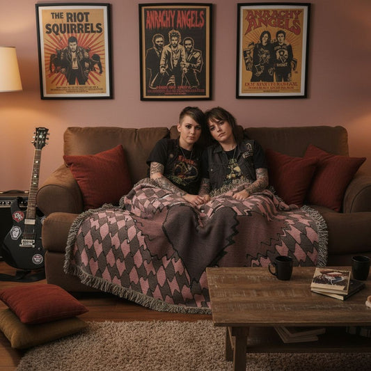 Two alt style people sitting on a couch, cuddled under a woven blanket with a dark distorted heart with wings. 